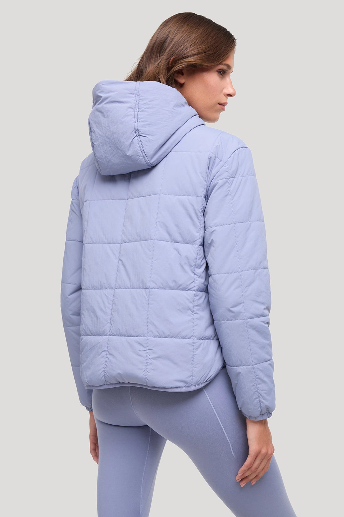 A woman wearing a light periwinkle blue quilted puffer jacket with a hood, shown from the back and side against a solid light gray background.