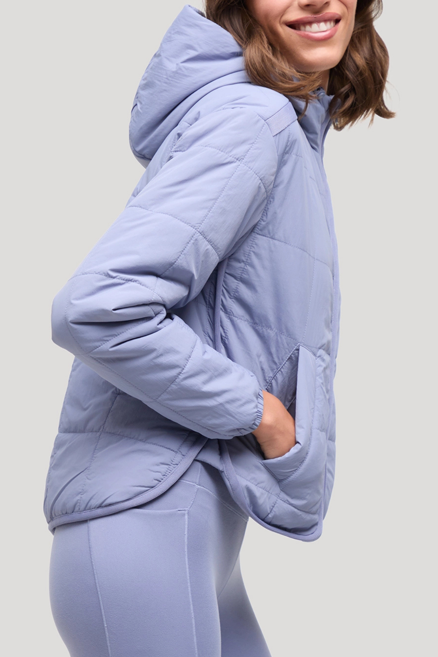 A woman wearing a light periwinkle blue quilted puffer jacket with a matching pair of leggings, shown from a side profile against a solid light gray background.