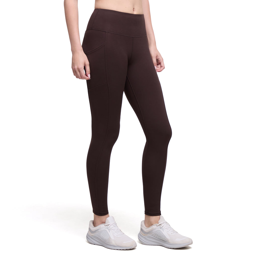 A model wearing dark brown high-waisted leggings with side pockets and white athletic sneakers, shown from the waist down on a white studio background.