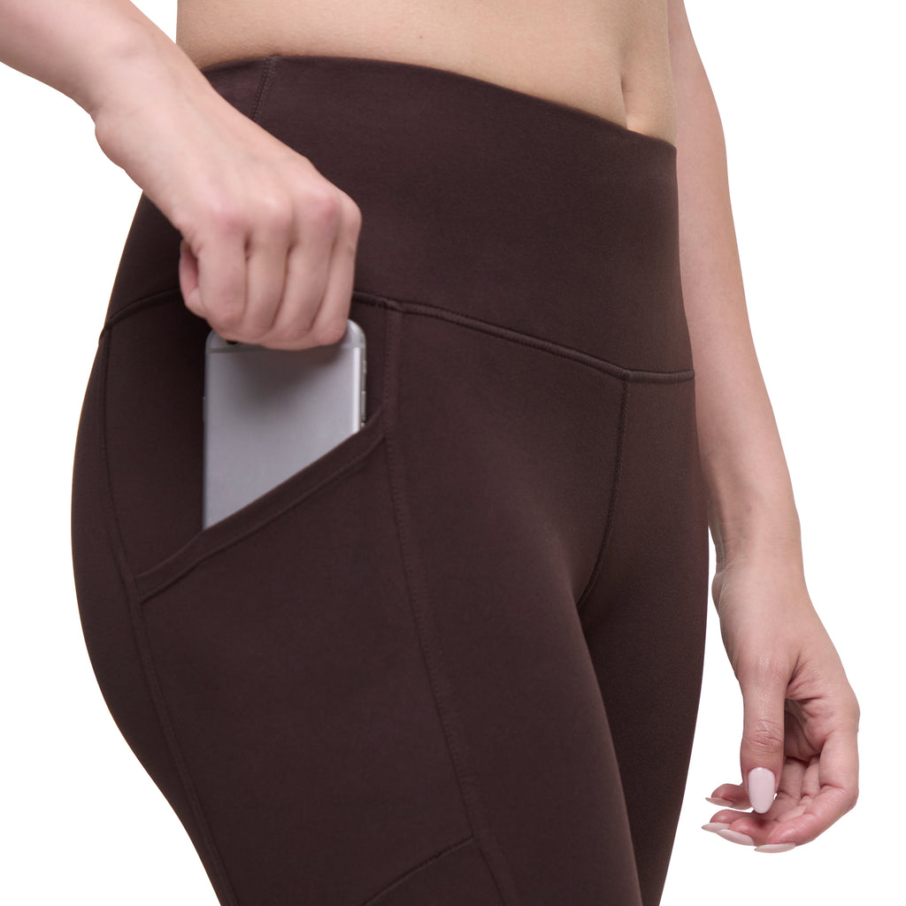 A close-up of a person wearing dark brown high-waisted leggings with a side pocket, shown holding a silver smartphone inside the pocket against a white background.