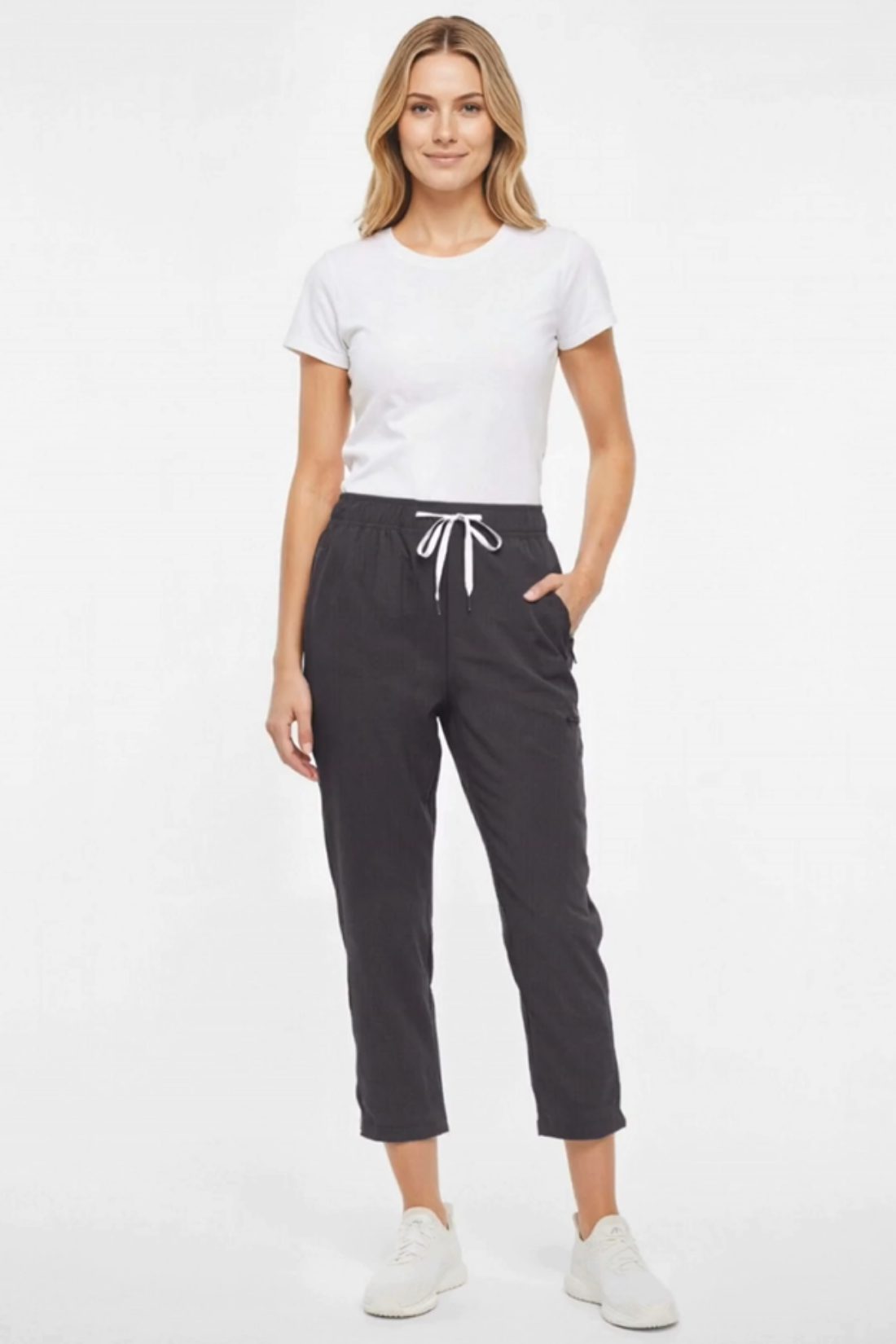 A woman modeling a white short-sleeve crewneck t-shirt and dark charcoal gray cropped jogger pants with a white drawstring, paired with white sneakers, against a plain white studio background.