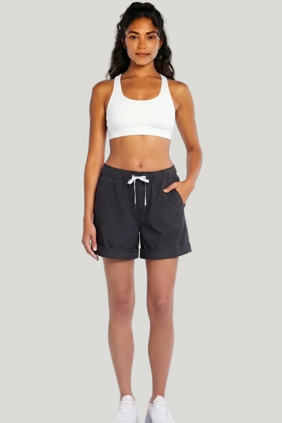 A woman modeling dark charcoal gray drawstring shorts and a white sports bra against a light gray studio background.