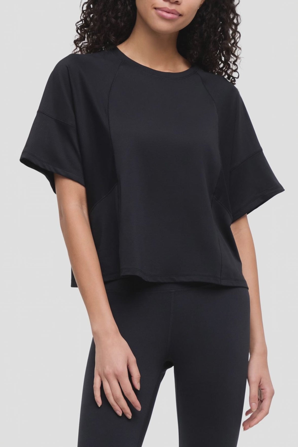 Video of a woman wearing a black short-sleeve crop top with raglan sleeves and black leggings, photographed against a light gray studio background. 