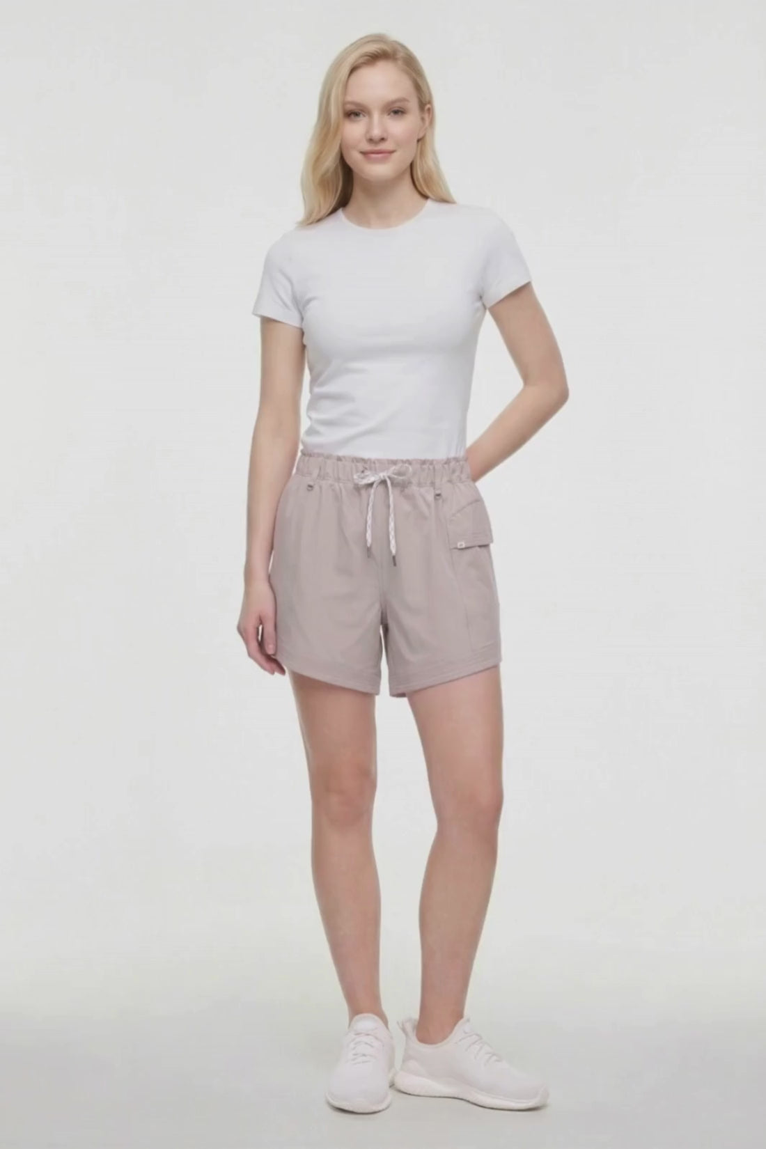 Video view of woman wearing beige shorts with white drawstring waist and side pocket and white sneakers on a white background. 