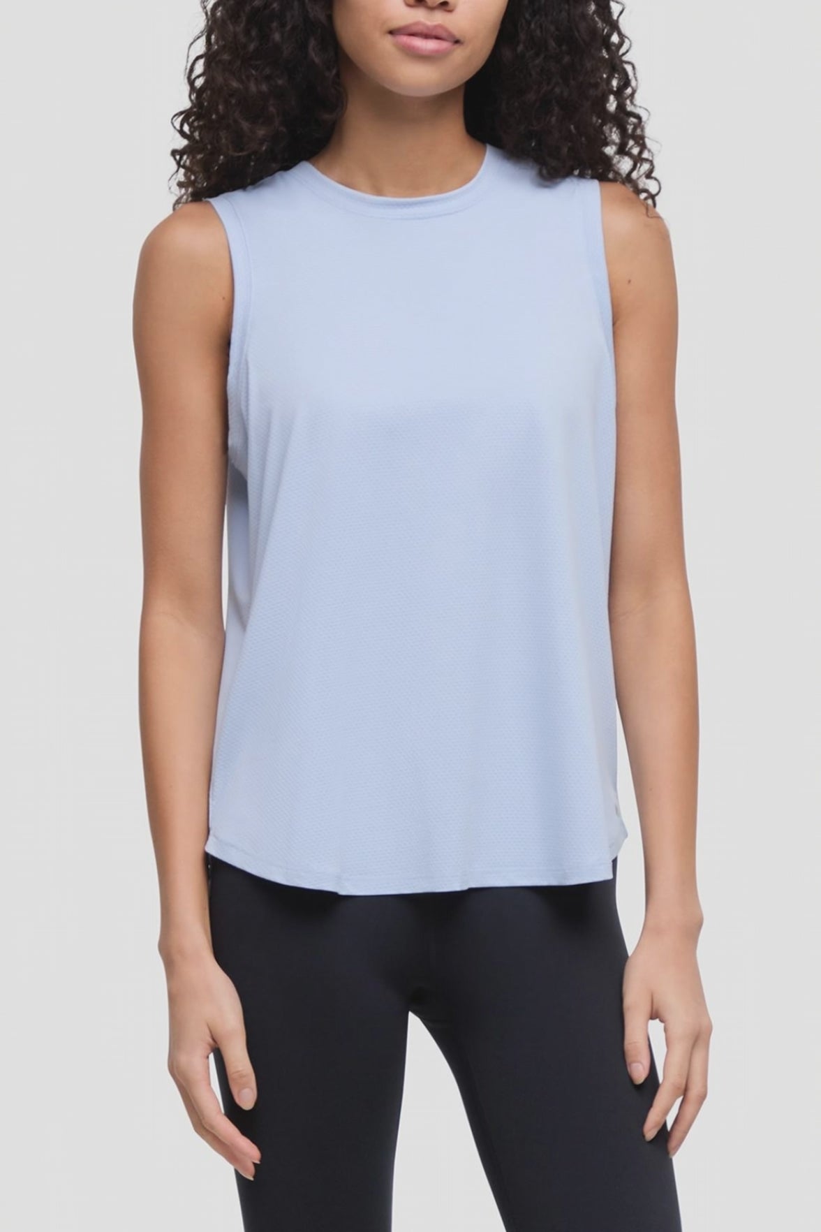 Video of a woman wearing a light blue sleeveless crewneck tank top and black leggings, shown from the waist up against a plain light gray studio background. 