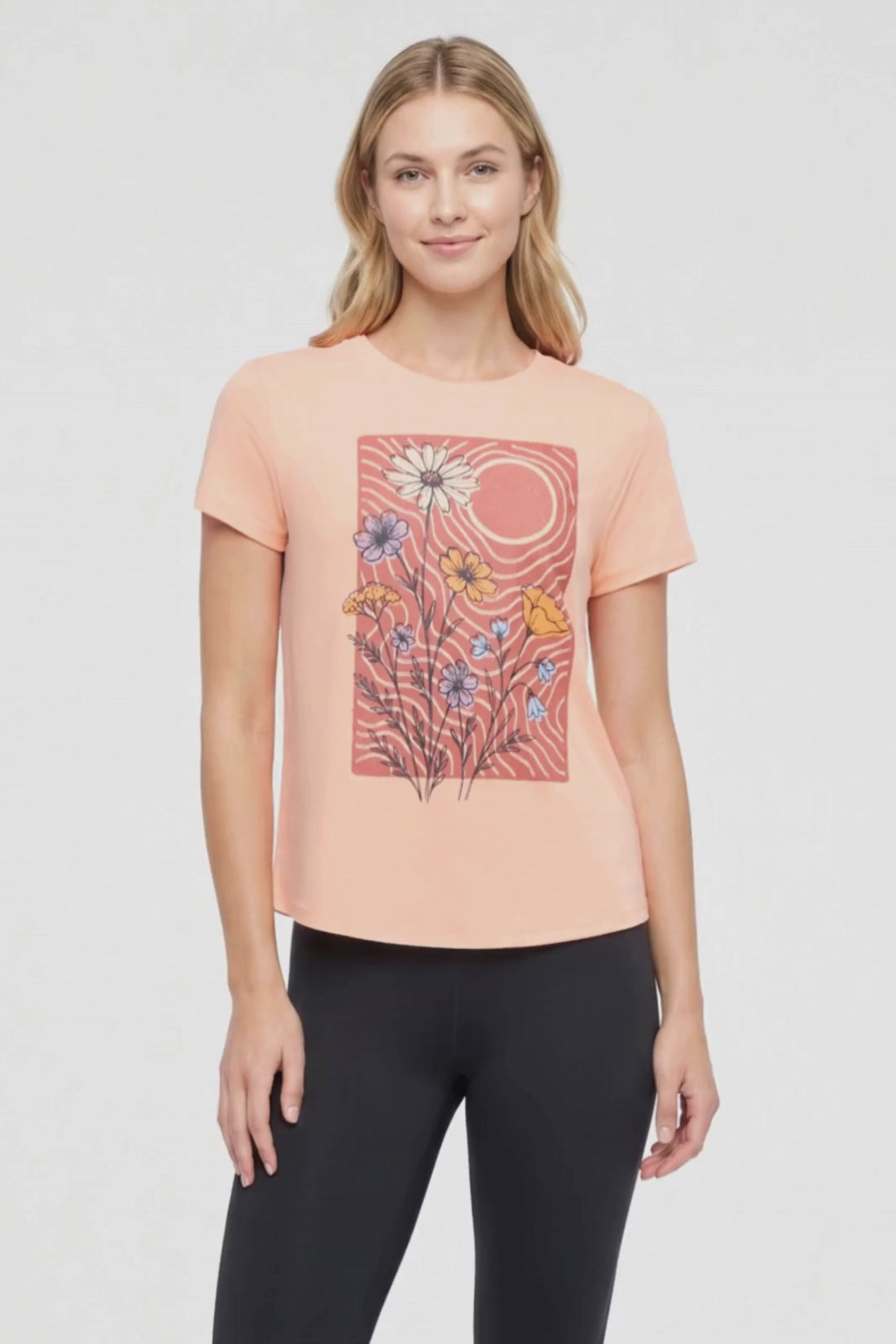 Video view of woman wearing a peach t-shirt with floral graphic on a light gray background. 