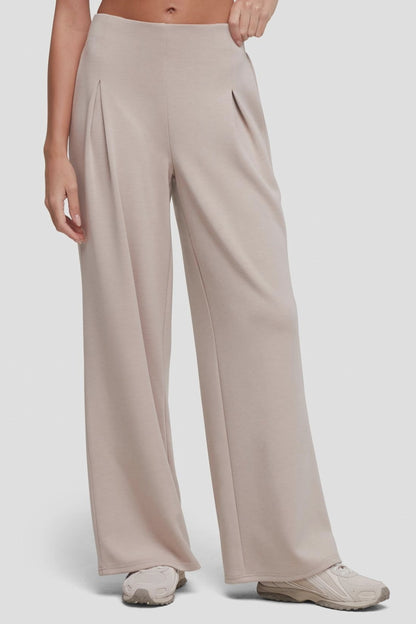 Video of A model wearing high-waisted, wide-leg pleated trousers in a light taupe-beige color with front pleat detail, paired with light-colored sneakers, against a plain light gray studio background. 