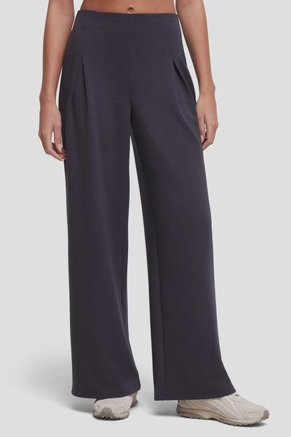 Video of a model wearing high-waisted, wide-leg trousers in a dark charcoal gray color with front pleat details, paired with beige sneakers, against a light gray studio background. 