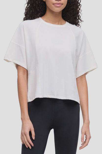 Video of a woman wearing a white short-sleeve crop top with wide sleeves and black leggings, shown from the shoulders to the upper thighs against a plain light background.  