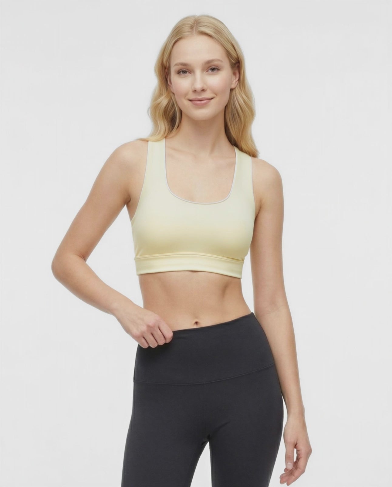 Video view of woman wearing a light yellow sports bra with white trim and black leggings on a white background. 