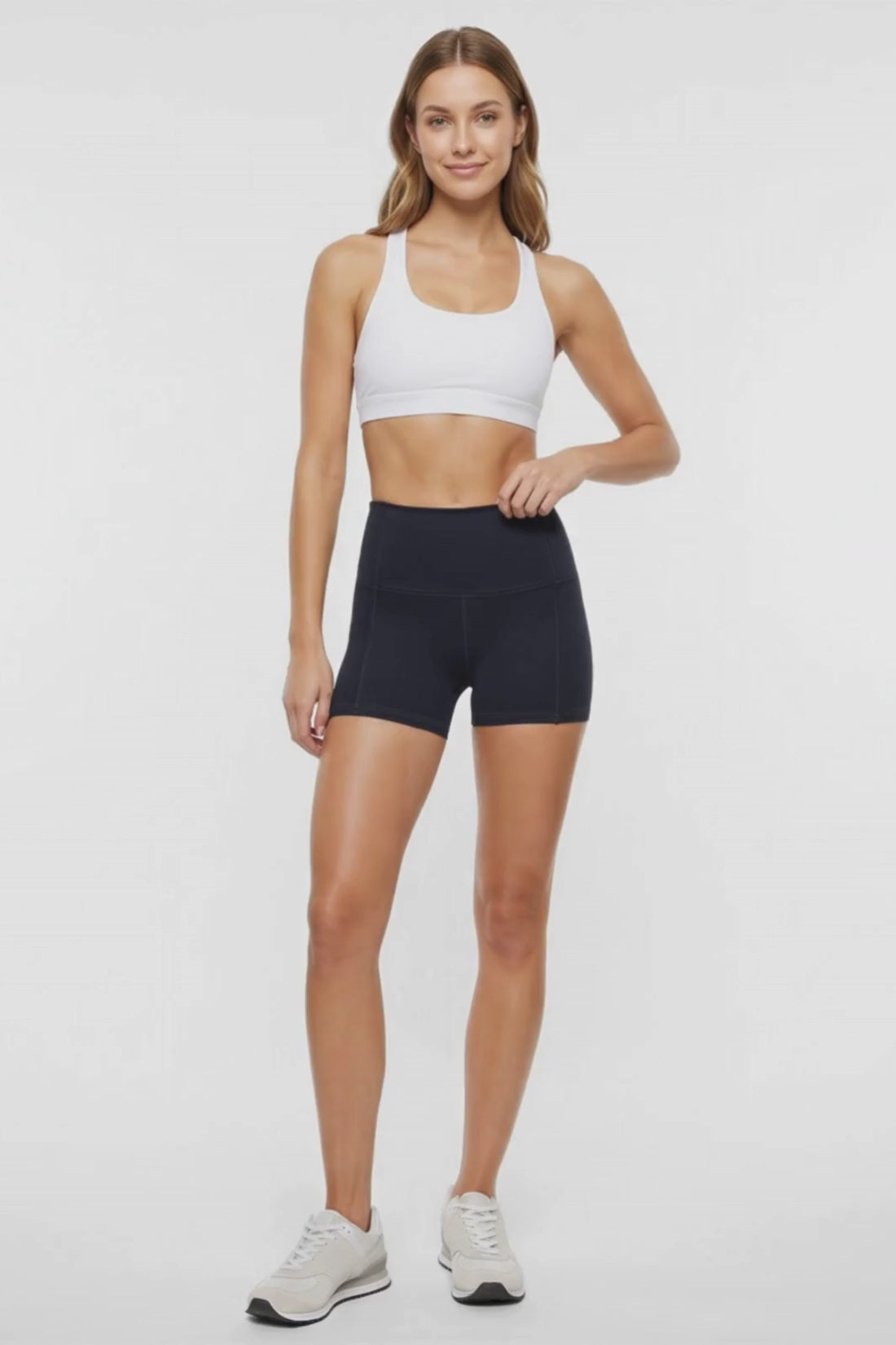 Video view of woman wearing black athletic shorts, white sports bra and white sneakers on a light background. 