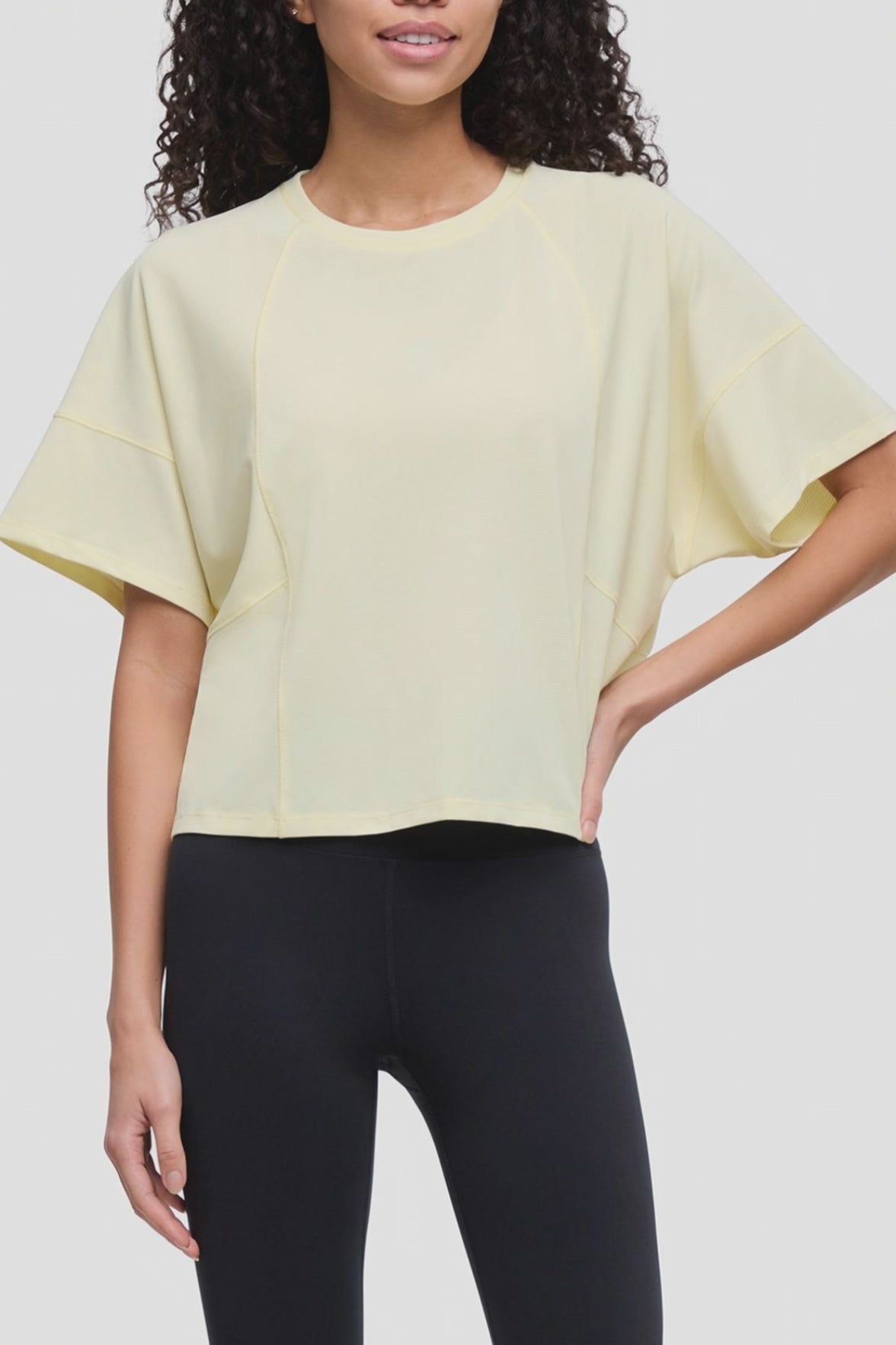 Video of a woman wearing a pale yellow short-sleeve crop top with wide draped sleeves and black leggings, shown against a light gray studio background. #color_tender-yellow